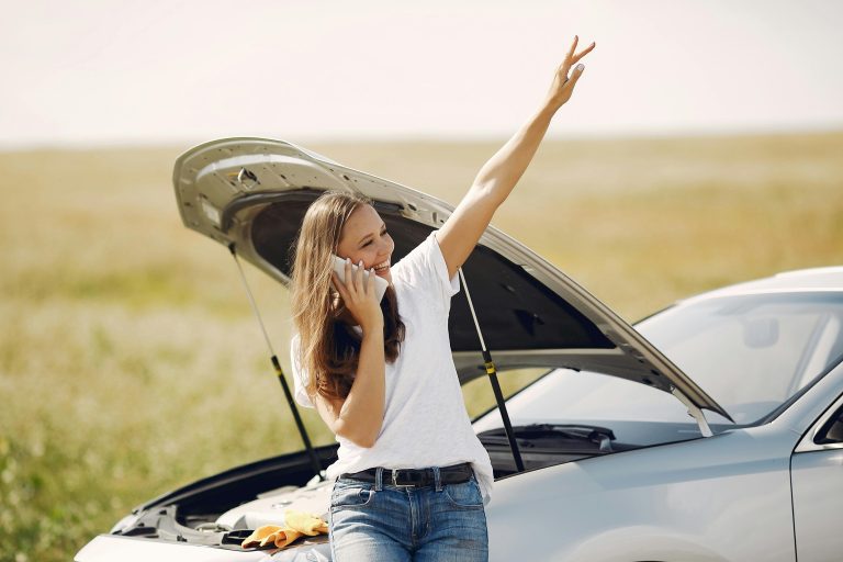 Positive woman using smartphone near broken automobile in countryside during car travel
